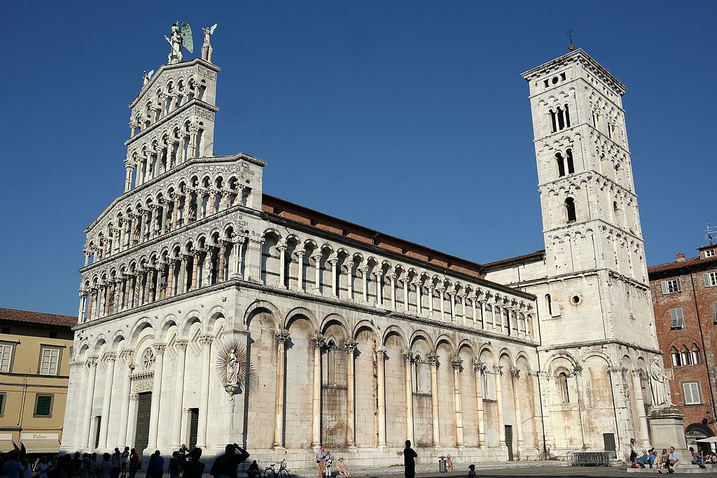 Chiesa di San Michele in foro con il campanile. La parte bassa ha arcate cieche per tutto il perimetro e nella parte più alta una fila di loggette. La facciata ha un profilo a a vela, è decorata con schiere di loggette sovrapposte e sormontate dalla statua di San Michele che sconfigge il drago e due angeli che suonano. Su un angolo della facciata cè un altare pensile con la statua della Madonna con bambino.