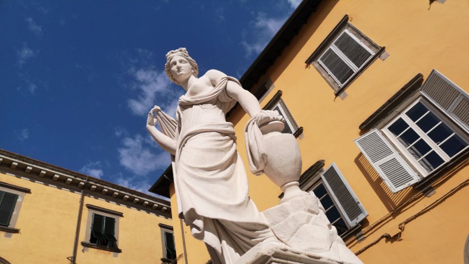 Statua neoclassica della fontana della Naiade, in piazza San Salvatore. Una figura femminile in calcare bianco, con una veste drappeggiata, capelli raccolti e un'anfora. Alle sue spalle i palazzi di Lucca.