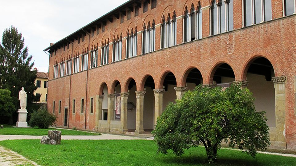 Facciata della villa Guinigi. un volume basso e lungo in mattoni. Nella parte bassa. al centro c'è un atrio con colonnato in pietra serena. Nella parte alta una specie di galleria formata da finestre a trifora. sulla destea la statua del carlaccio