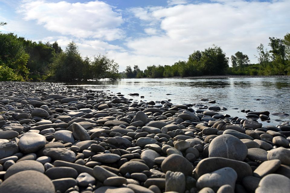 Primo piano di sassi su una spiaggetta sul fiume Serchio che scorre tranquillo tra la vegetazione e sotto un cielo azzurro con nuvole estive.