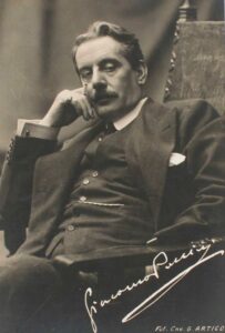 fotografia di Giacomo Puccini seduto su una poltrona. Indossa un completo elegante con panciotto, fazzoletto nel taschino, gemelli ai polsini della camicia. in mano ha una sigaretta. Nell'angolo in basso a destra c'è la sua firma