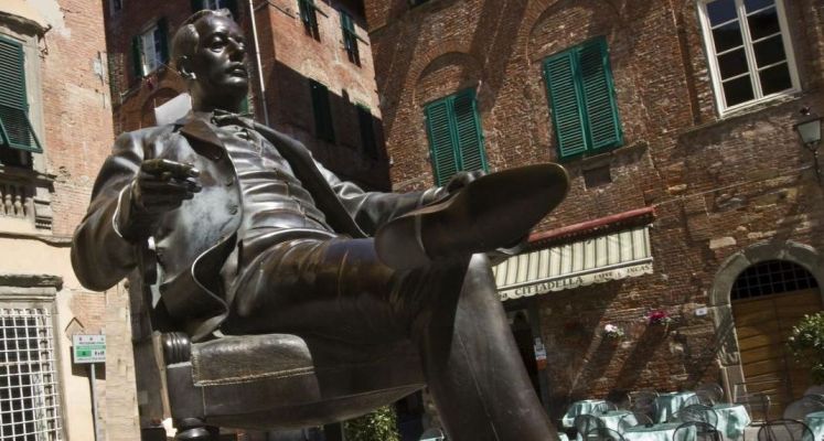 Statua di bronzo Giacomo Puccini in piazza Cittadella. Puccini è seduto su una poltroncina con una gamba accavallata e un sigaro in mano. Sullo sfondo case in mattoni e l'edificio che ospita il museo casa natale