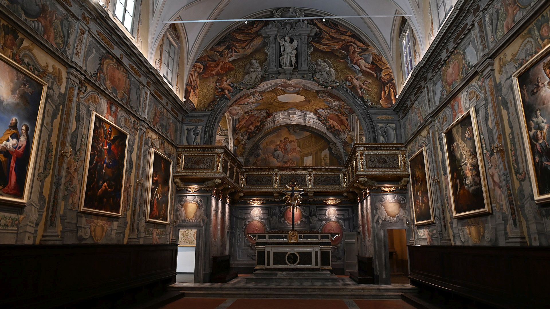 Interno dell'Oratorio degli Angeli Custodi con affresco a trompe l'oeil raffigurante architetture barocche, stucchi dorati e putti che reggono festoni di fiori. Alle pareti laterali sono appesi grandi dipinti.
