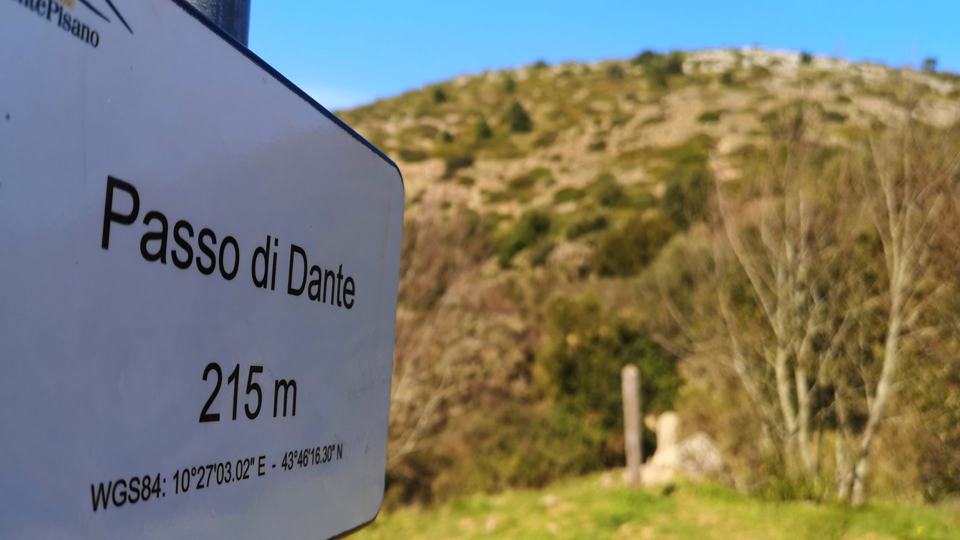 un cartello segnaletico indica il passo di Dante a 215 metri di altezza sul livello del mare. Sullo sfondo il Monte Pisano con la vegetazione bassa.