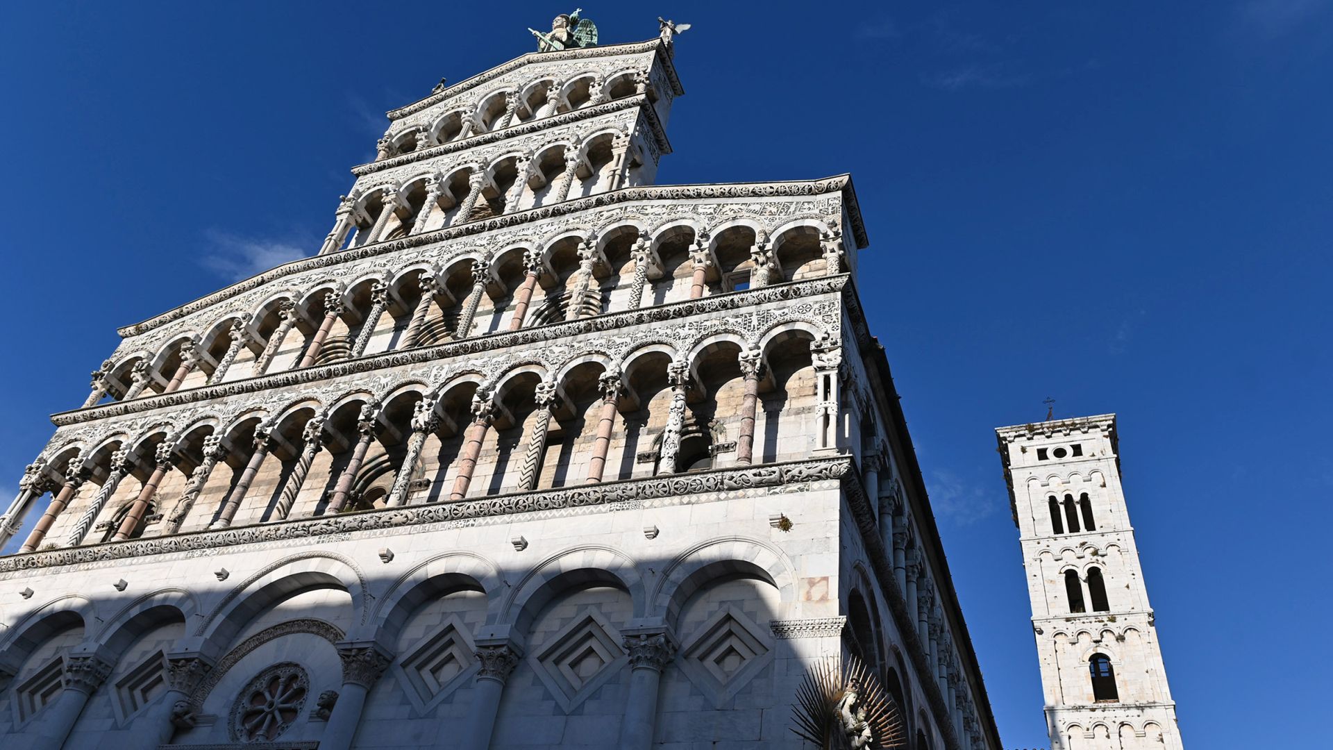 facciata della chiesa di San Michele con il campanile. Al piano terra della facciata c'è una fila di arcate cieche in cui si aprono tre portera. Sopra ci sono 4 file di loggette con colonne earchitravi fittamente decorati. In cima alla facciata c'è la statua di San Michele Arcangelo