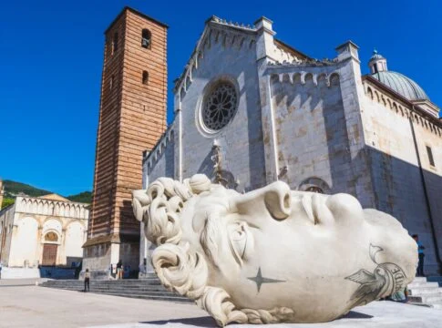 Copia della testa del David di Michelangelo in piazza del Duomo a Pietrasanta. Sul fondo il duomo gotico con campanile in mattoni e la chiesa di Sant'Agostino dalla facciata rettagolare con tre archi ciechi nella parte bassa.