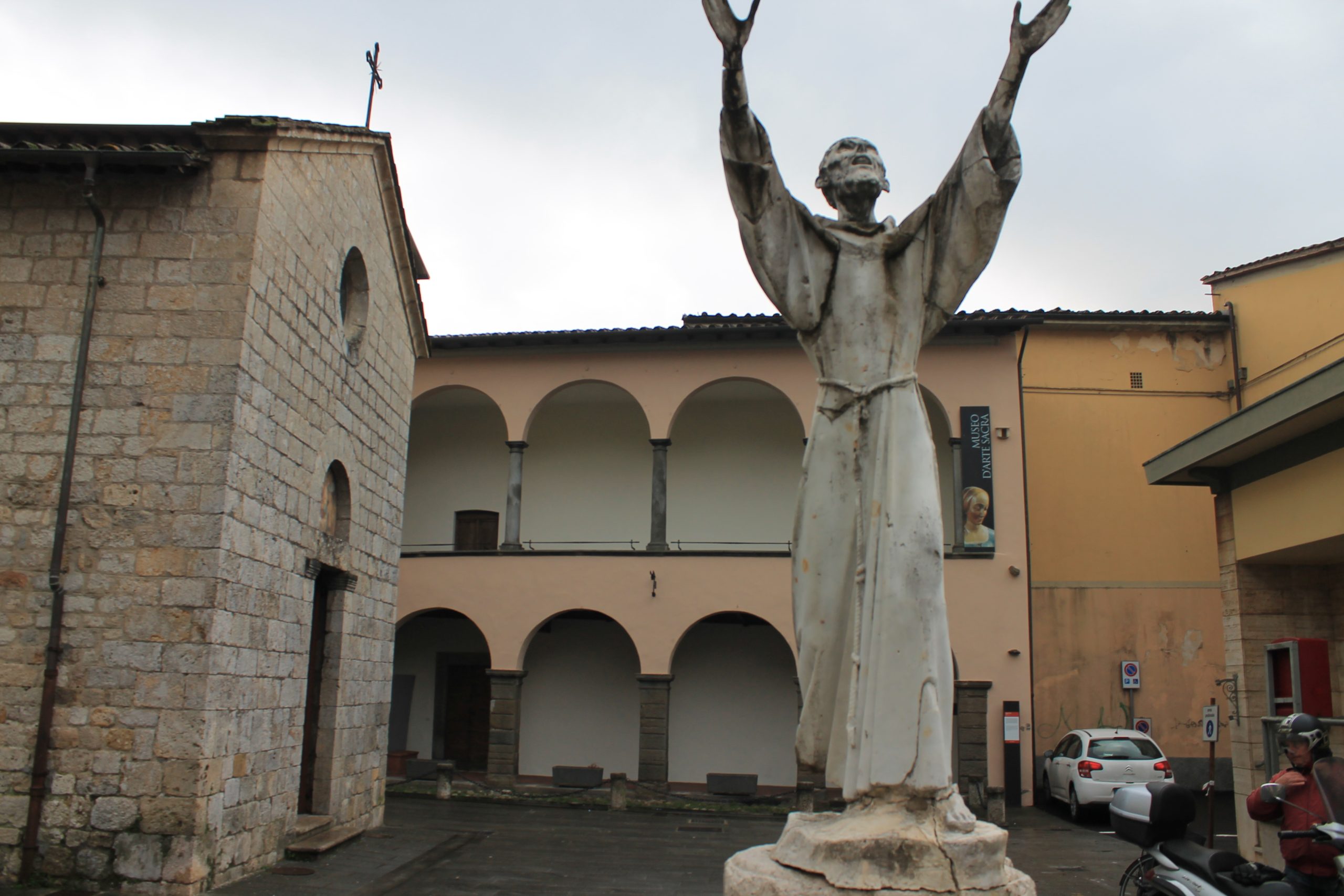 Statua di San Francesco con le mani al cielo di fronte al Museo di Arti Sacre di Camaiore e alla piccola chiesa di San Francesco.