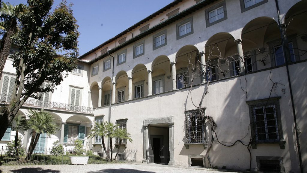 Faciata interna del Museo Nazionale di Palazzo Mansi su 4 piani. Al piano terra un portale in pietra seerena e tre finestre. Su un lato un piccolo giardinetto. Al piano nobile una loggia con archi e colonne in pietra serena su cui si arrampica una vite selvatica spoglia. Al piano superiore finestre rettangolari con cornici di pietra serena come all'utlimo piano, arretrato.