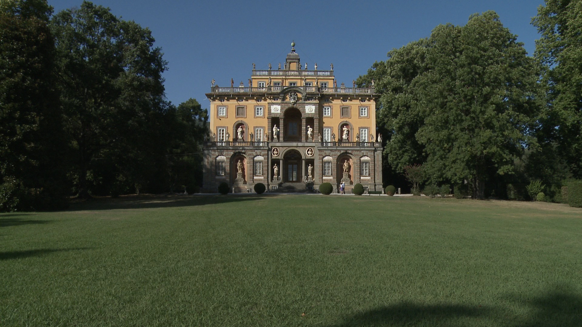Facciata della Villa Torrigiani di Camigliano. Un intonaco color ocra su cui spiccano decorazioni architettoniche in pietra serena e statue in calcare bianco. Di fonte alla villa un prato all'inglese e di lato grandi alberi frondosi
