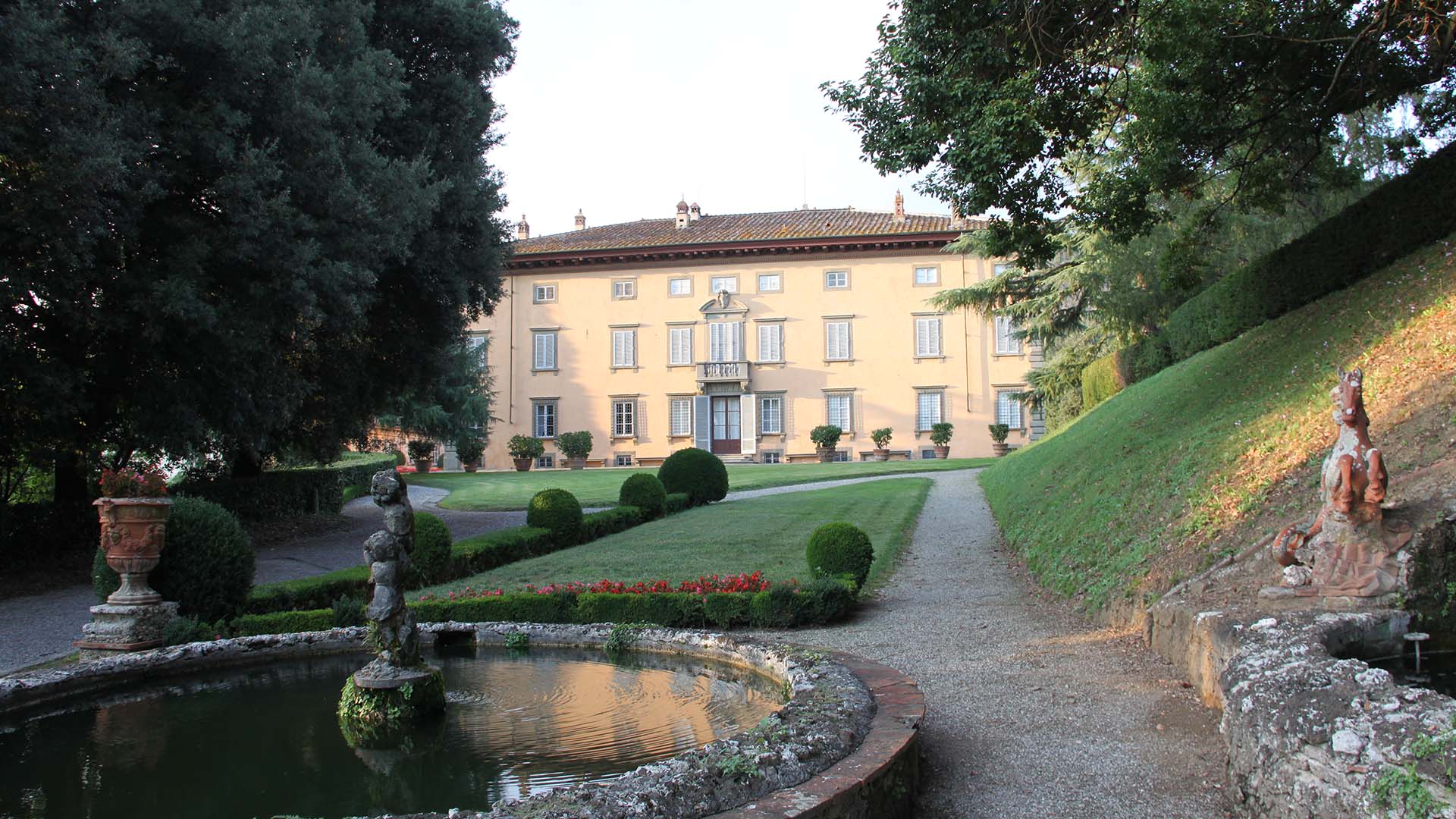 Facciata sud di Villa Oliva in stile neoclassico e giardino. Al centro della facciata della villa c'è un portale di ingresso sormontato da un terrazzino. In primo piano una fontana con statua di un putto, il prato all'inglese con cespugli di bosso e fiori.