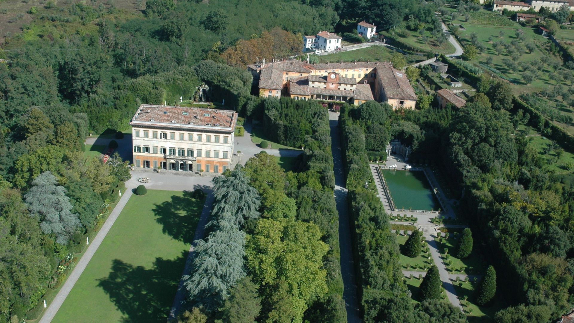 panoramica dall'alto del giardino di Villa Reale di Marlia immersa nel verde. Sulla sinistra la villa, Davanti alla villa il prato all'inglese e sul retro il teatro dell'acqua con statue e fontane, al centro il viale di alloro che prota alla palazzina dell'Orologio e sulla sinistra la peschiera con il giardino dei limoni. Intorno c'è la campagna con olivi.