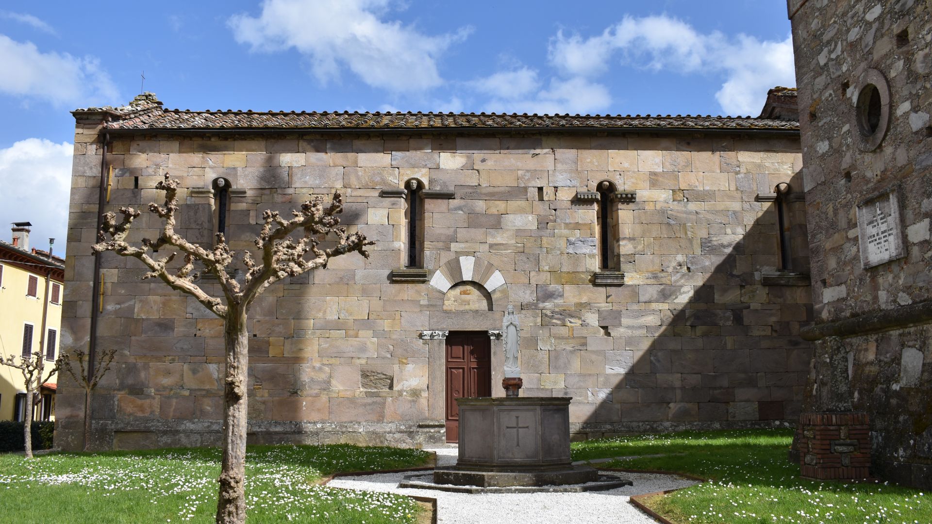 lato sud della pieve di San Leonardo in treponzio con tre finestre strette e lunghe sormontate da piccole teste scolpite. Di fonte all'edificio c'è un giardinetto con un albero e un fonte battesimale