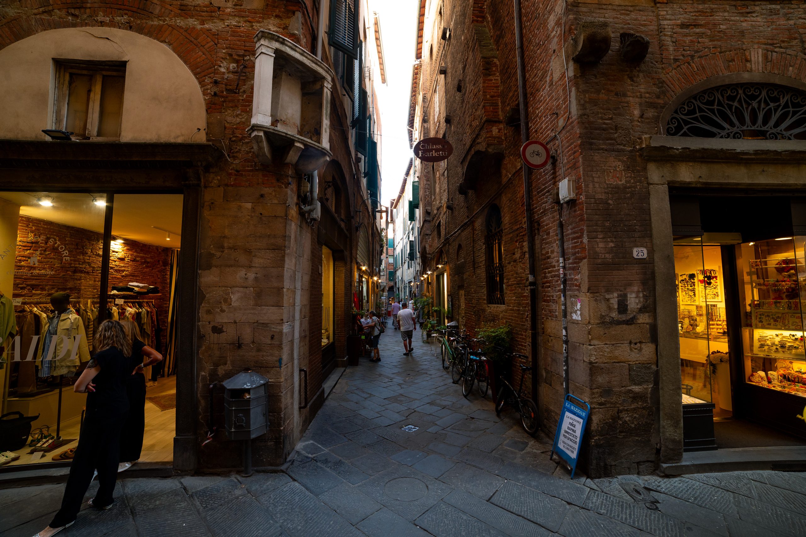 Chiasso Barletti nel centro storico di Lucca. Una piccola strada con vetrine illuminate, persone che camminano e biciclette apoggiate al muso
