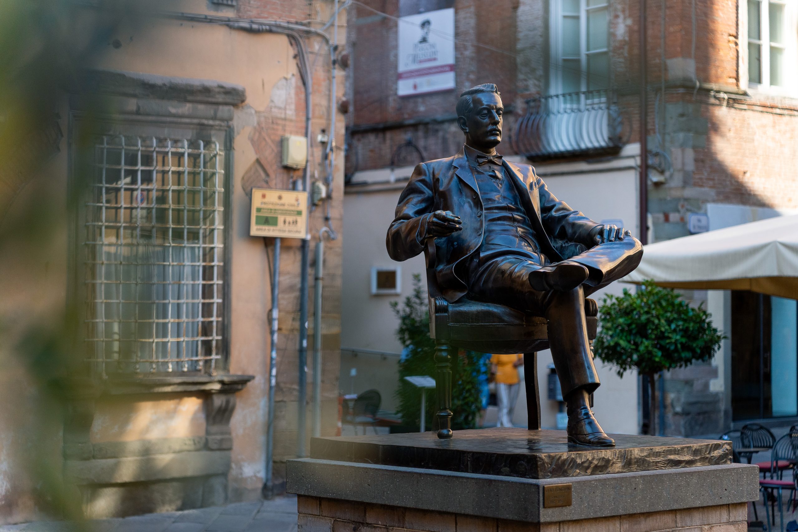 Statua di Giacomo Puccini che fuma seduto in poltrona in piazza della cittadella. Sullo sfondo la casa natale