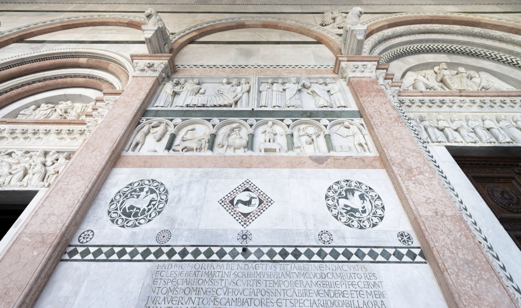 Particolare della facciata della cattedrale di San Martino con bassorilievi e intarsi in marmo di diversi colori. Nei bassorilievi sono raffigurati i mesi dell'anno come contadini. Luglio batte il grano, agosto raccoglie i frutti, Sttembre pigia l'uva nel tino, ottobre toglie il vino dalla botte, novembre ara i campi con due buoi, dicembre ammazza il maiale. Sopra ci sono i bassorilievi con le storie di San Martino. A sinistra San Martino guarisce un indemoniato sdraiato nel suo letto e accudito da tre donne. Dalla sua bocca esce un diavoletto. Due frati aiutano san Martino.