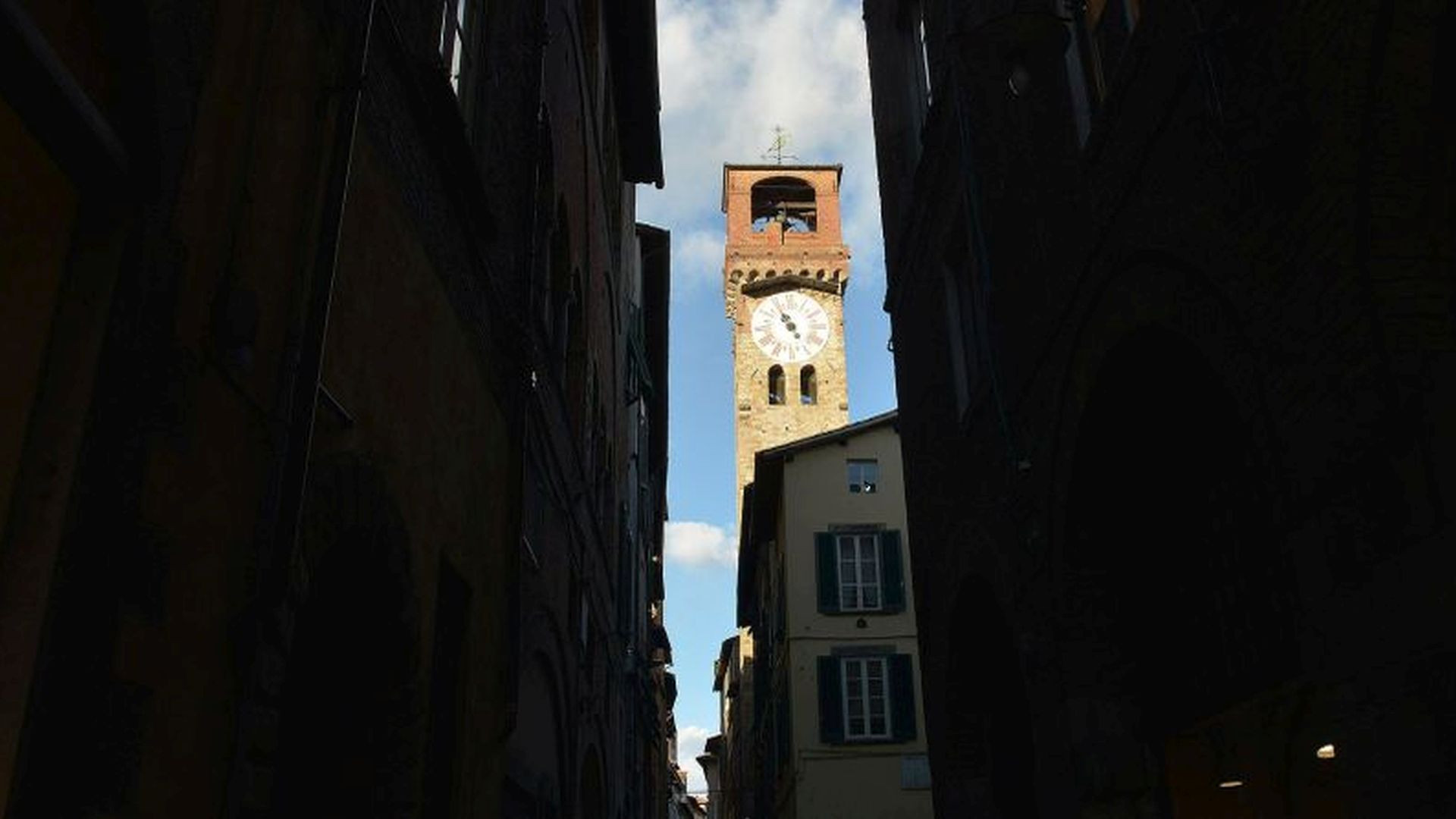 Dal canto d'Arco (incrocio di via Fillungo con via Roma), tra gli alti edifici controluce si staglia il cielo azzurro con la Torre delle Ore. Sulla torre il quadrante del grande l'orologio setecentesco e in cima il campanario coperto con la banderuola che reca la scritta libertas