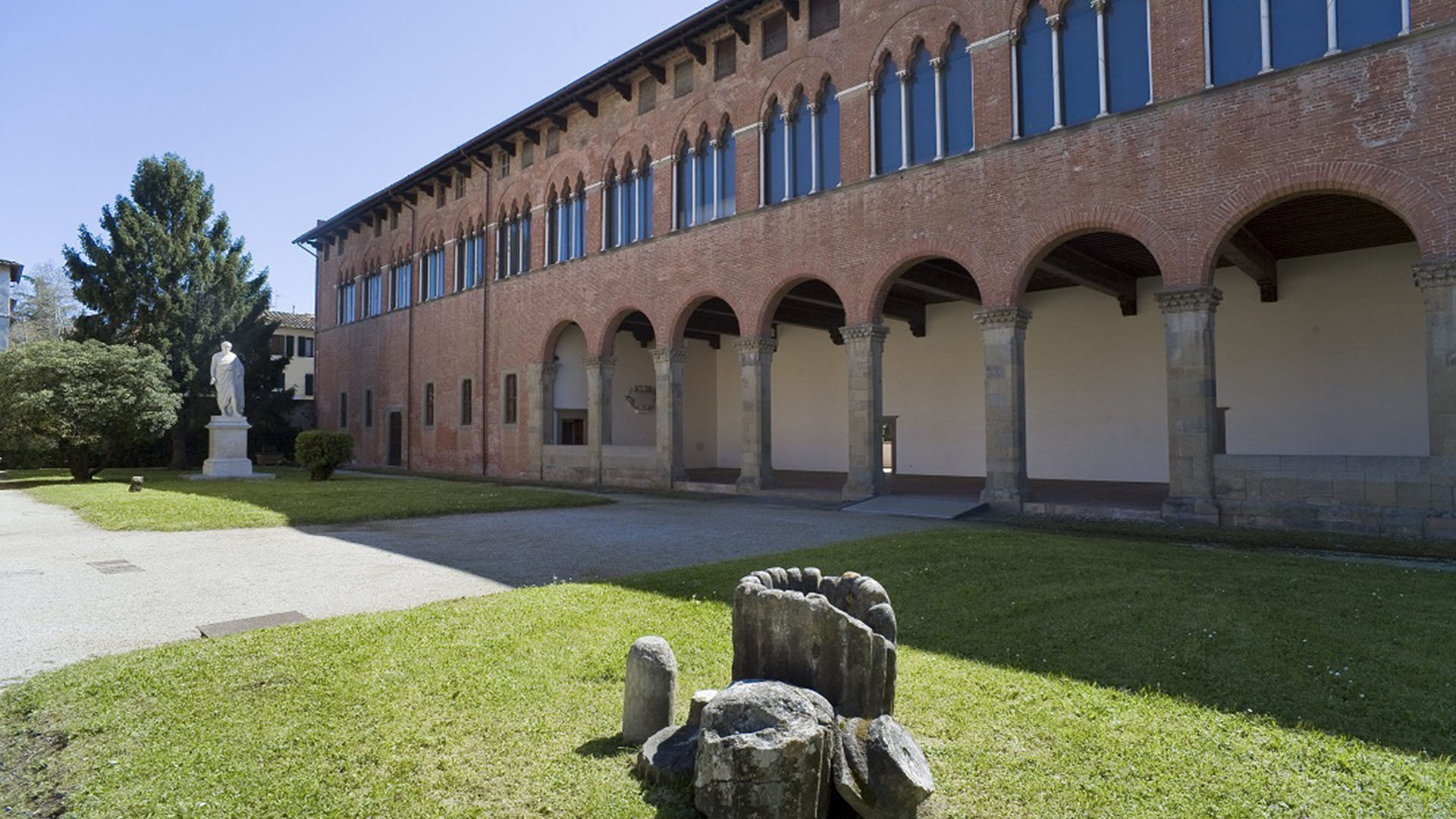 facciata del Museo Nazionale di Villa Guinigi. Un edificio piuttosto largo, a due piani. Al Piano terra un grande atrio con archi e lesene di pietra serena. Al piano superiore finestre continue a trifora in stile gotico. Sopra le piccole finestre quadrate della soffitta. Sul prato di fronte resti romani, su un lato in fondo, la statua di Carlo IV