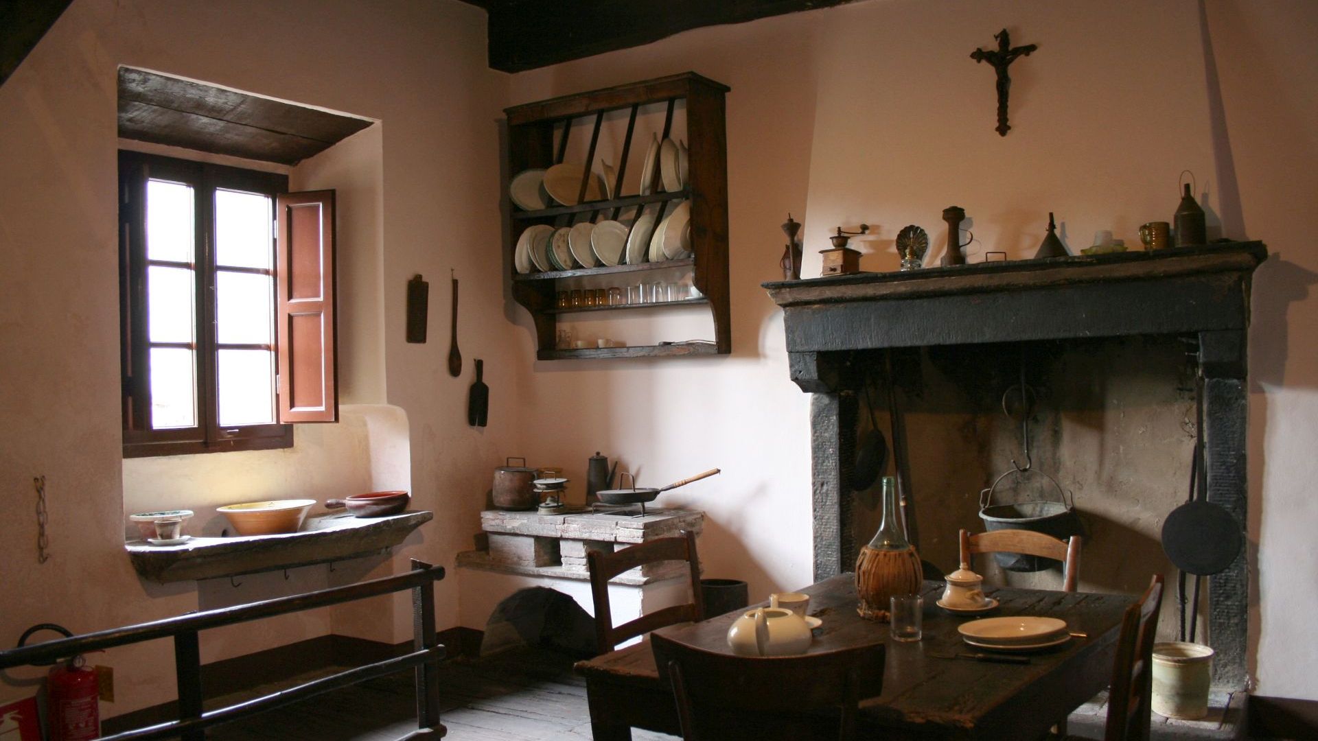Cucina di casa Puccini a Celle dei Puccini. Una stanza luminosa arredata semplicemente, con un tavolo apparecchiato, una cucina a legna in muratura, un grande caminetto con un paiolo, una piattaia in legno con piatti e un lavandino in pietra sotto la finestra.