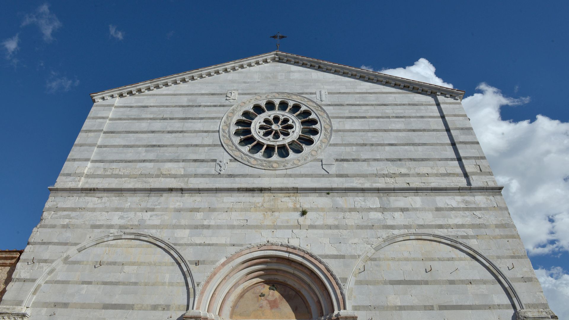 Scorcio dal basso della parte alta della facciata della chiesa di San Francesco. La facciata è molto lineare con profilo a capanna e decorazione a fasce alterne di marmo bianco e verde. Nella parte alta c'è un granderosone.
