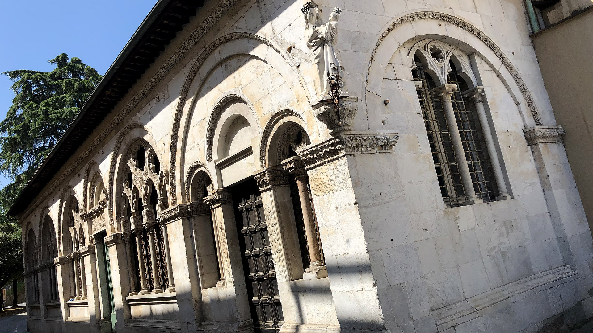 Spicolo esterno della chiesa della Rosa. Sullo spicolo c'è una statua della madonnna con bambino e una rosa su una mensola. I due hanno una corona di ferro battuto. Sul fianco destro dello spigolo, sotto un arco decorato con bassorilievi, si apre una bifora con arco gotico e un oculo. Sul lato sinistro una serie di archi decorati con bassorilievi. si Nel più vicino si apre una porta fiancheggiata da finestre a bifora. Poi seguono grandi finestre a quadrifora con archi gotici e oculi gotici.
