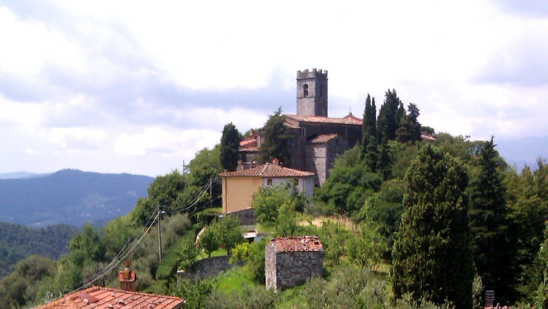 Panorama del paese di Deccio di Brancoli. In cima ad un colle boscoso, alcune case in pietra e la sagoma della chiesa con il campanile a merli quadrati.