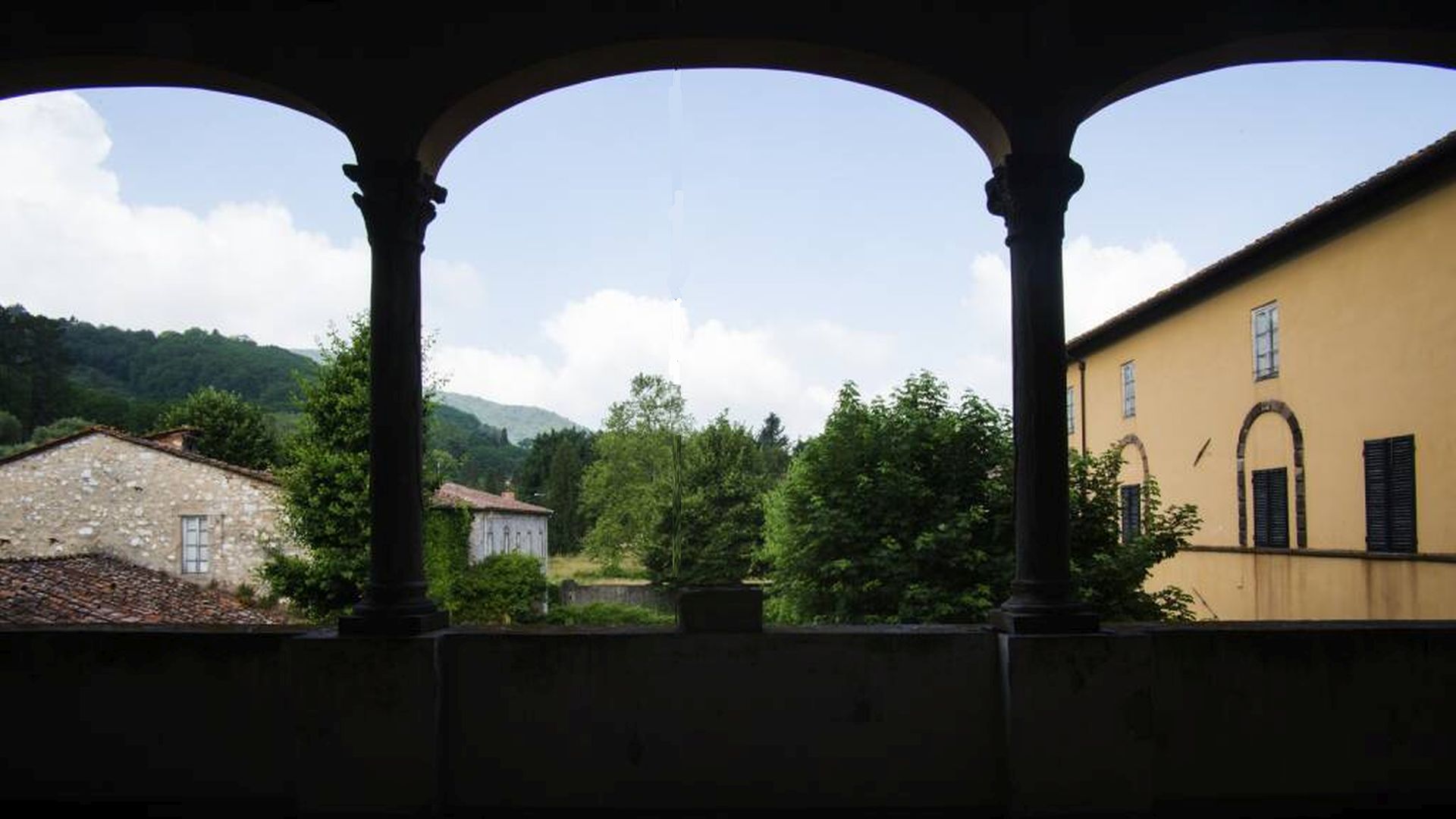 panorama dal loggiato del primo piano del monastero della Visitazione di VIcopelago. Oltre gli archi, sulla destra c'è l'edificio del monastero, di fronte due case rurali e alberi. Sullo sfondo le colline.