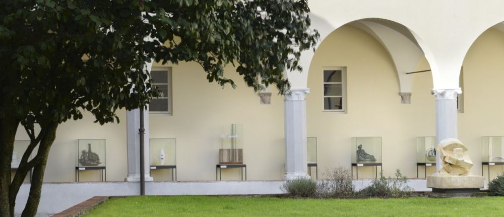 Chiostro di San MIcheletto. Sotto il loggiato ci sono delle sculture contemporanne in teche di vetro. In primo piano c'è la chioma di un albero e un piccolo giardino con una scultura.