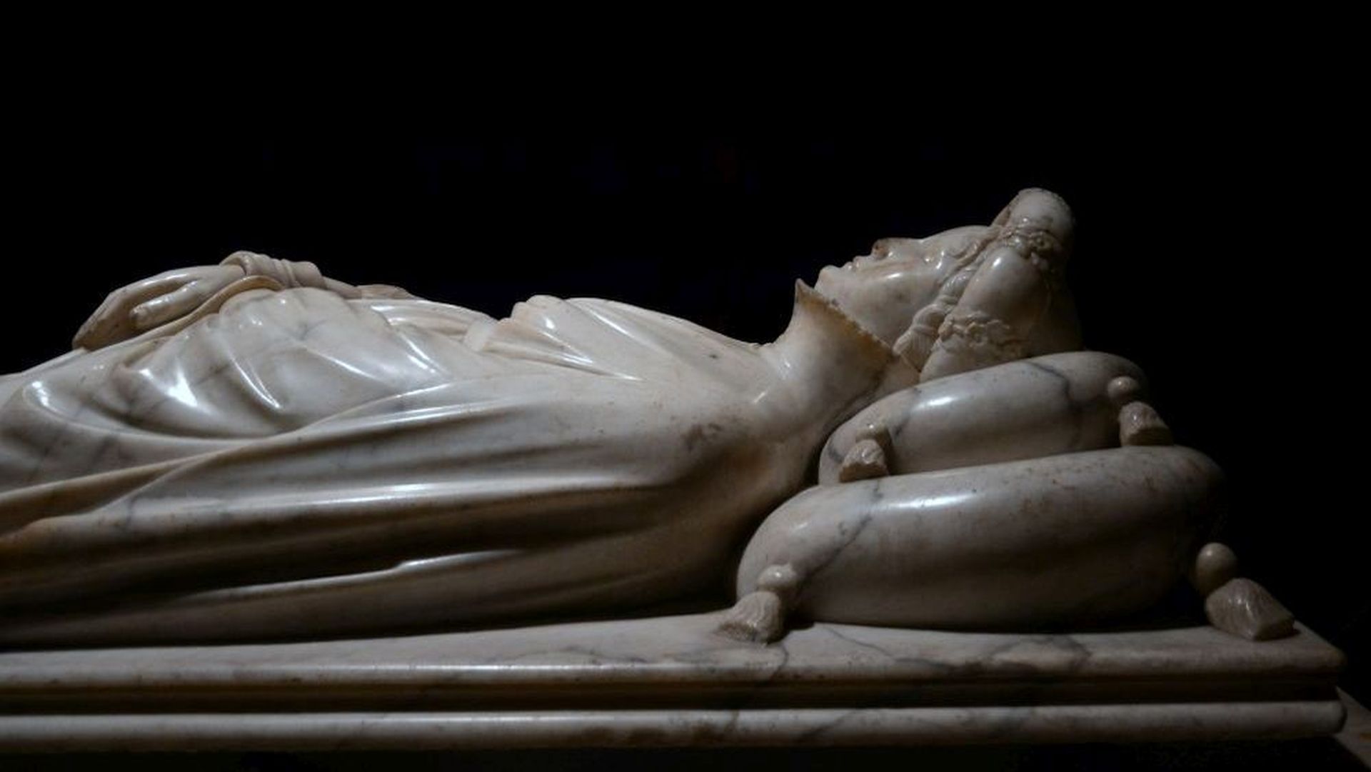 Monumento funebre di Ilaria del Carretto di profilo. Ilaria riposa sul suo letto con la testa appoggiata su due cuscini, le mani giunte sul ventre. Indossa un ricco abito di velluto e una corona di fiori.