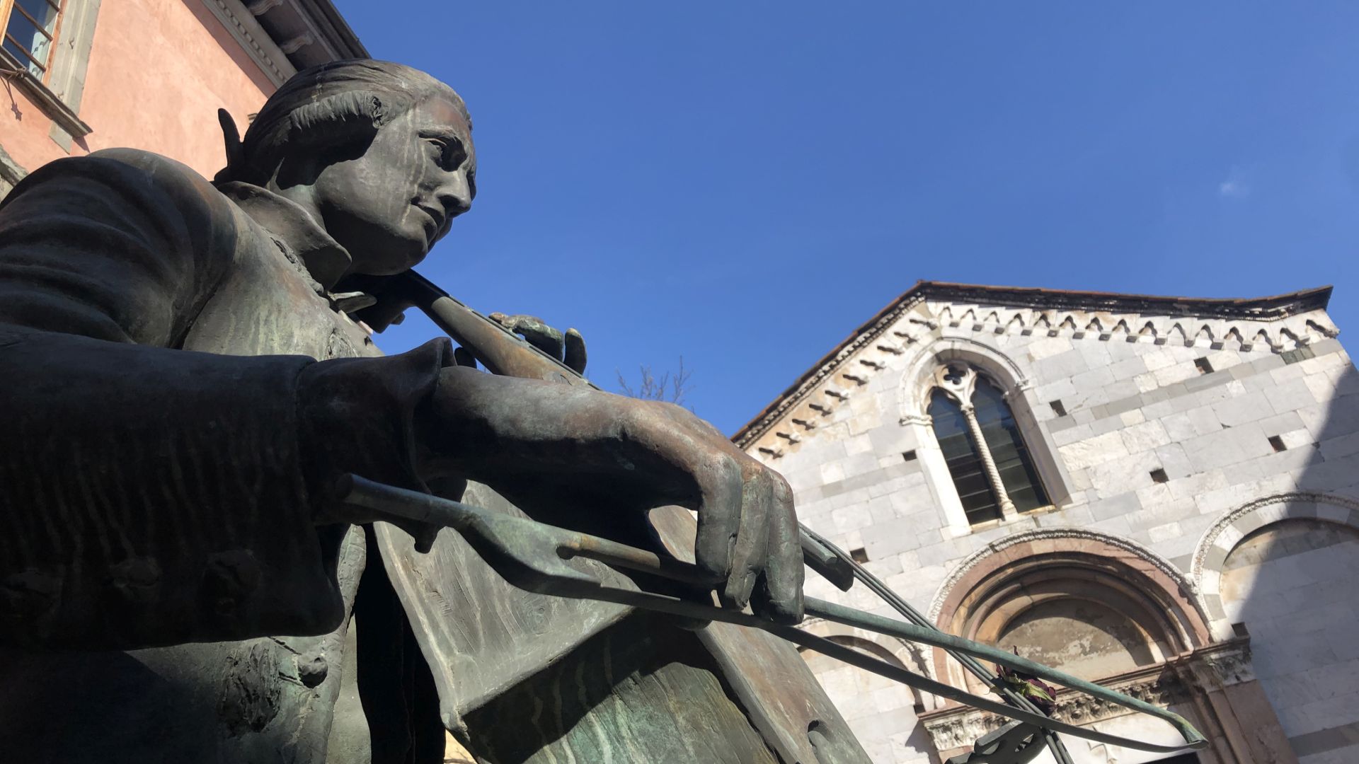 Scultura in bronzo di Luigi Boccherini in abiti settecenteschi che suona il viloncello davanti alla chiesa gotica di Santa GIulia
