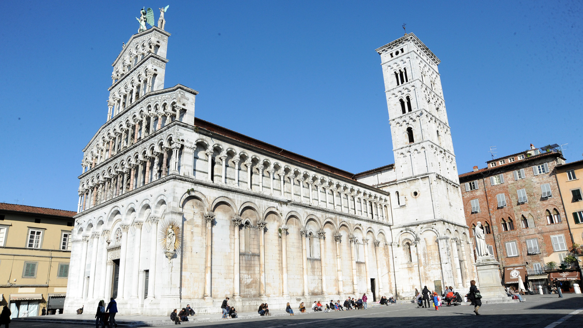 Piazza San MIchele con persone sedute sugli scalini. La chiesa ha una decorazione con arcate cieche nella parte più bassa per tutto il perimetro e 4 loggette nella parte superiore della facciata a vela. IN cima alla faccaita la statua di San Michele arcangelo. Su uno spigolo della chiesa un altare pensile con la vergine con bambino