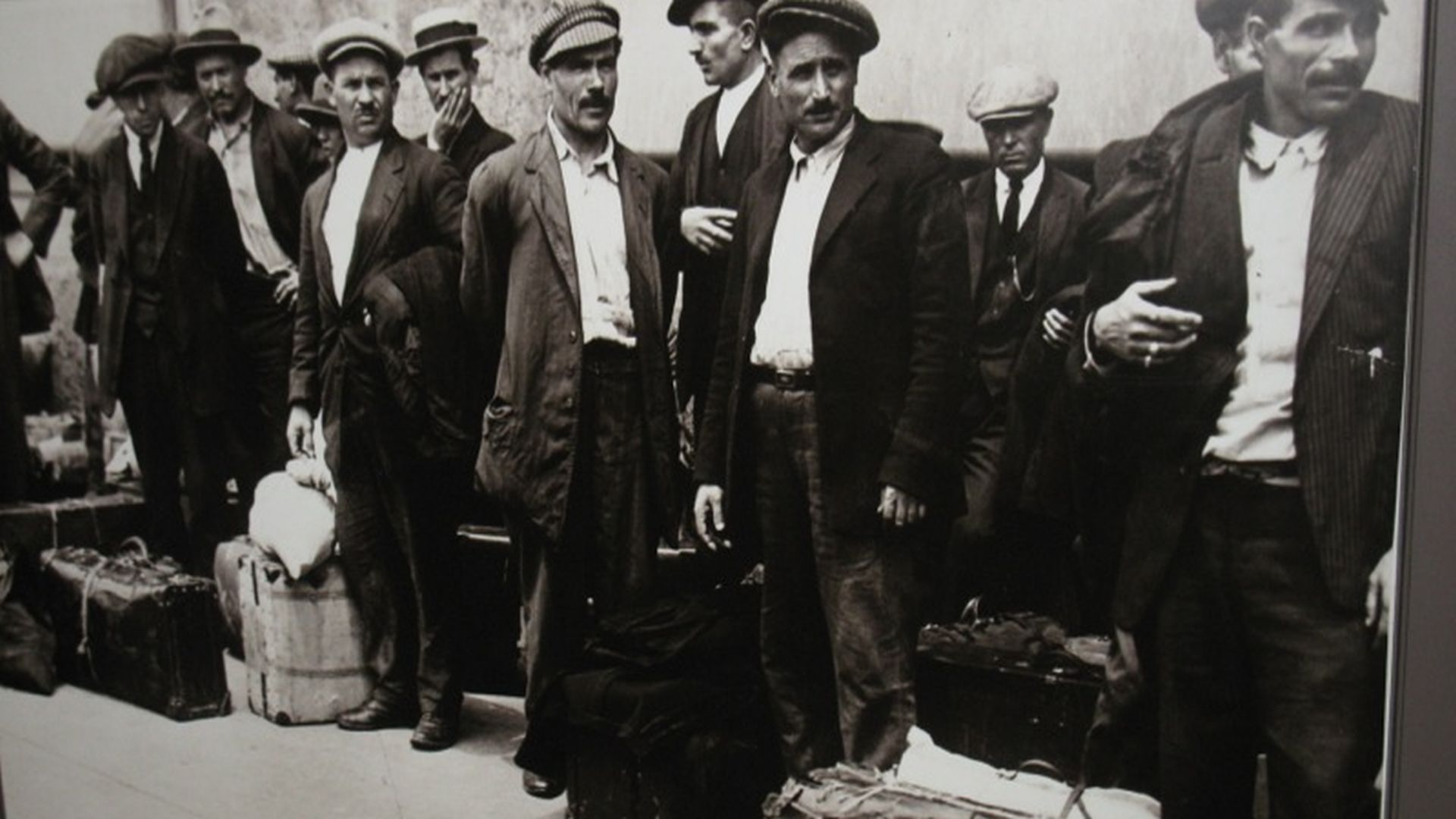 foto d'epoca con alcuni uomini in fila, in attesa di imbarcarsi. Hanno abiti consunti e fagotti e valigie legati con corde