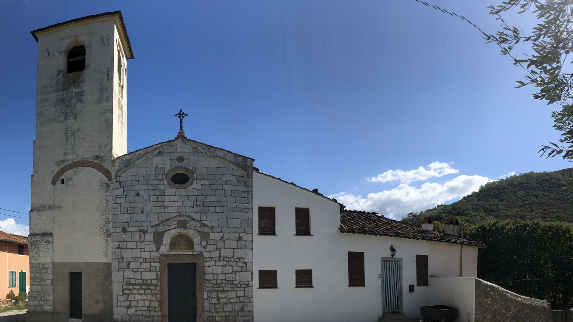 facciata della piccola chiesa di Meati tra le case e il campanile. La facciata è lineare con tetto a capanna e con l'unica apertura della porta sovrastata da un oculo. Sul fondo i boschi sui rilievi del Monte Pisano