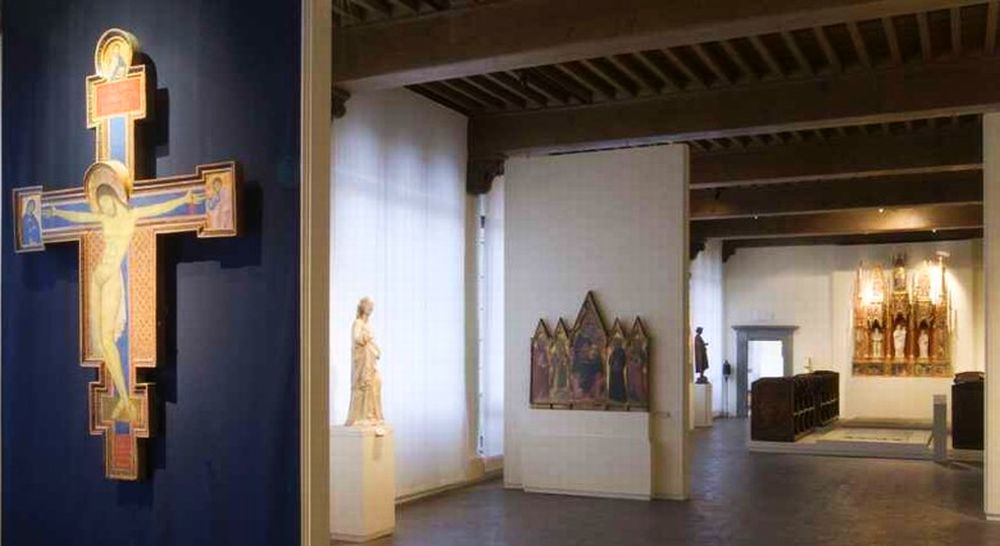 prospettiva di sale del Museo Nazionale di VIlla Guinigi. In primo piano unaparete su cui è appesa una croce dipinta duecentesca, segue una sala con una scultura di Madonna e un polittico raffigurante la vergine con bambino e quattro santi, sullo sfondo una sala con un'edicola gotica con statue della vergine con bambino e due santi. Ai suoi piedi una lapide