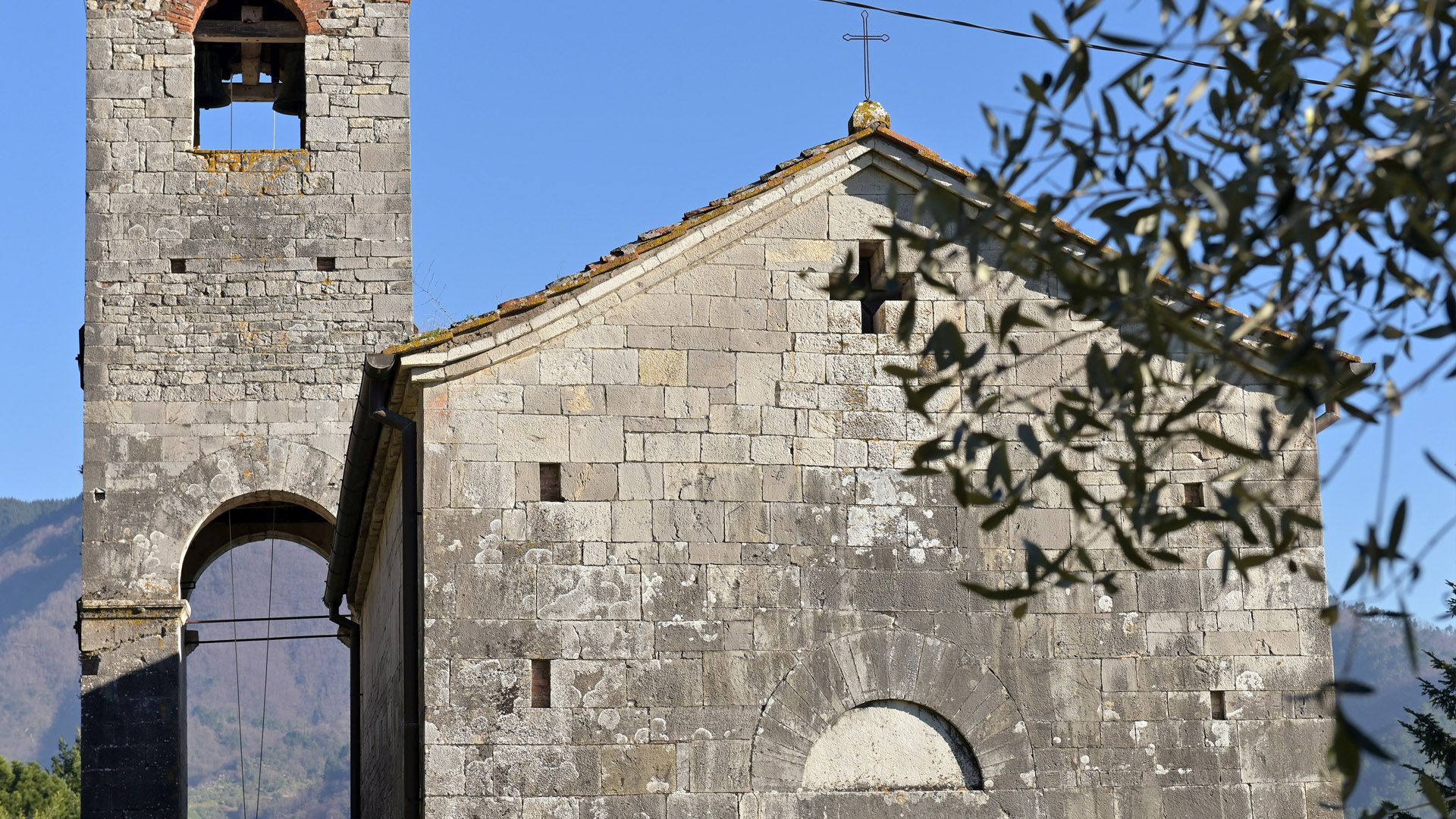 Particolare della parte alta della facciata e della pare centrle del campanile della pieve di San Lorenzo in Corte. La chiesa ha una faccaita semplice con tetto a capanna e una sola apertura centrale sormontata da una croce. Il campanile è in pietra, molto semplice ma con un passaggio nella aprte bassa e la cella campanaria coperta.