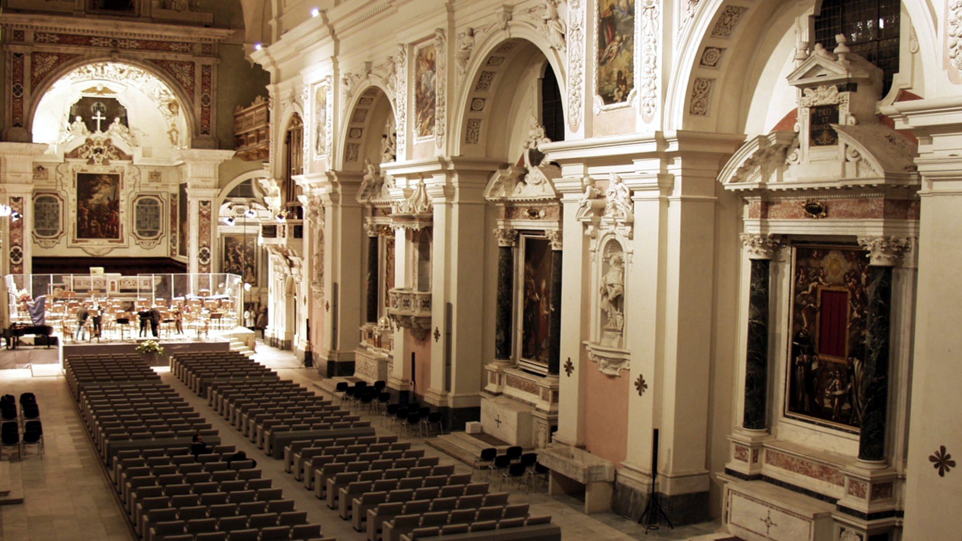 Interno della Auditorium Chiesa di San Romano dall'alto. Sul pavimento poltoncine per il pubblico. Sulla destra una parete riccamente decorata in stile manierista con sequensa di altari contenuti in grandi archi e divisi da lesene con decorazioni a stucco dipinti e statue. Sulla sinistra l'arco dell'altare maggiore, con l'altare su cui pende un grande quadro. Davanti all'altare un palco allestito per un'orchestra
