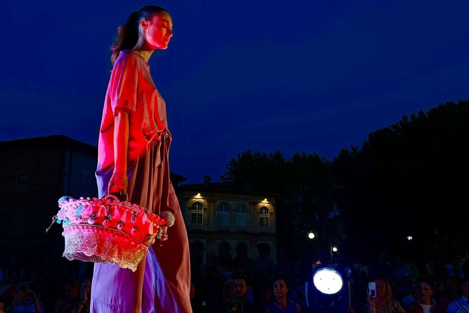 una sfilata serale di Lucca fashion weekend, In primo piano unamodella con un abito drappeggiato e una borsa etnica illuminata da una luce colorata.