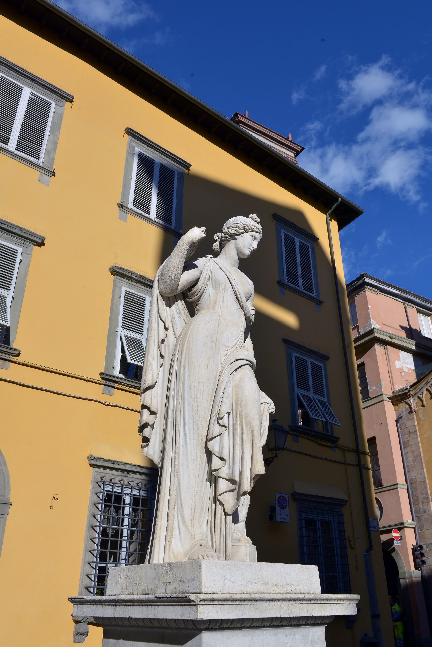 Statua neoclassica della fontana della Naiade, in piazza San Salvatore. Una figura femminile in calcare bianco, con una veste drappeggiata, capelli raccolti e un'anfora.. Alle sue spalle i palazzi di Lucca.
