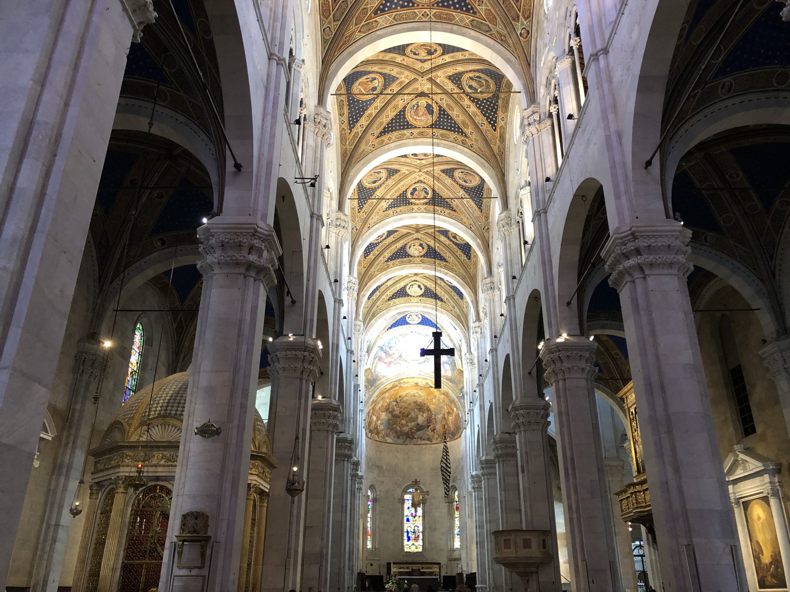 Veduta centrale della navata della cattedrale di Lucca con il soffitto dipinto a cielo stellato. Da un arco pendono una croce e una croce dentata. Nella natvata sinistra c'è il tempietto del Volto Santo