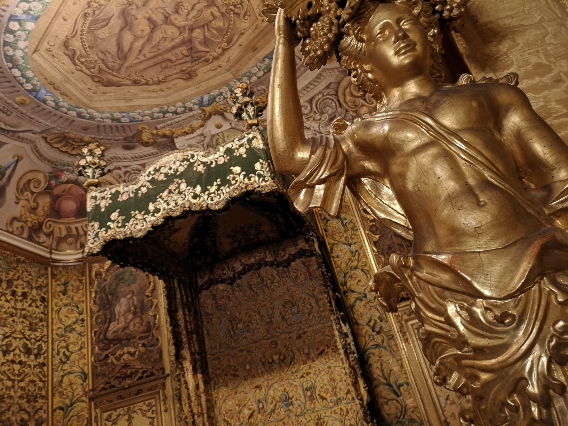 Alcova degli sposi di Palazzo Mansi. Sulla destra la statua dorata di un personaggio con frutta in testa. alle sue spalle l'alcova con il baldacchino ricamato in seta con fiori e frutti. Tappezzerie coordinate e soffitto affrescto con scene personaggi che riposano.