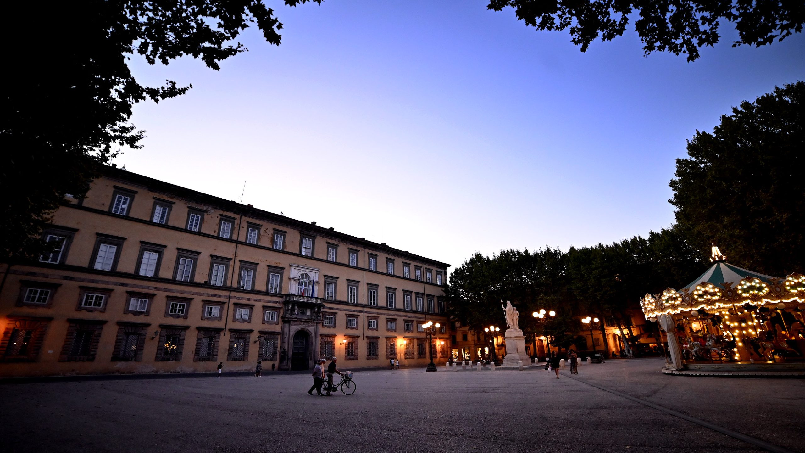 Piazza Napoleone di sera. Ci sono due persone che attraversano la piazza in bicicletta e, sulla sinistra, la giostra illuminata.