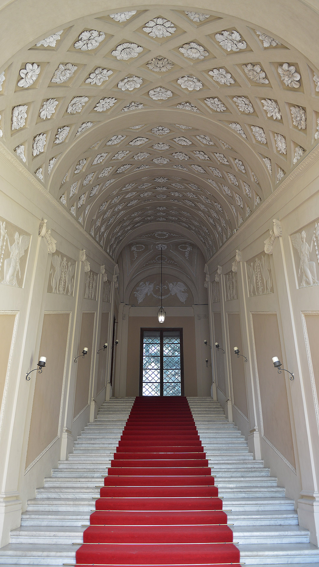 prospettiva dello scalone monumentale di Palazzo Ducale con tappeto rosso. La volta è a cassettoni decorati con fiori.