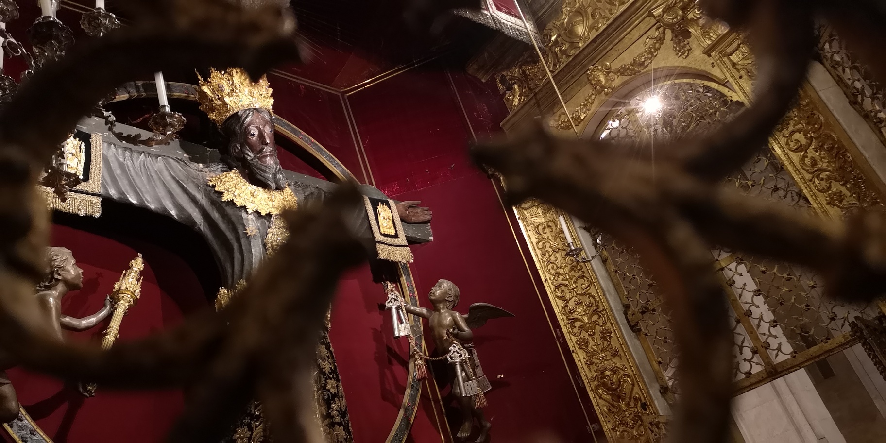 Particolare del Volto Santo prima del restauro attraverso la grata del tempietto. Il Volto Santo Indossa le vesti rituali delle festività, un collare e una corona d'oro, manicotti in velluto bordati di ricami dorati. due angeli ai lati della croce porgono lo scettro e le chiavi della città.