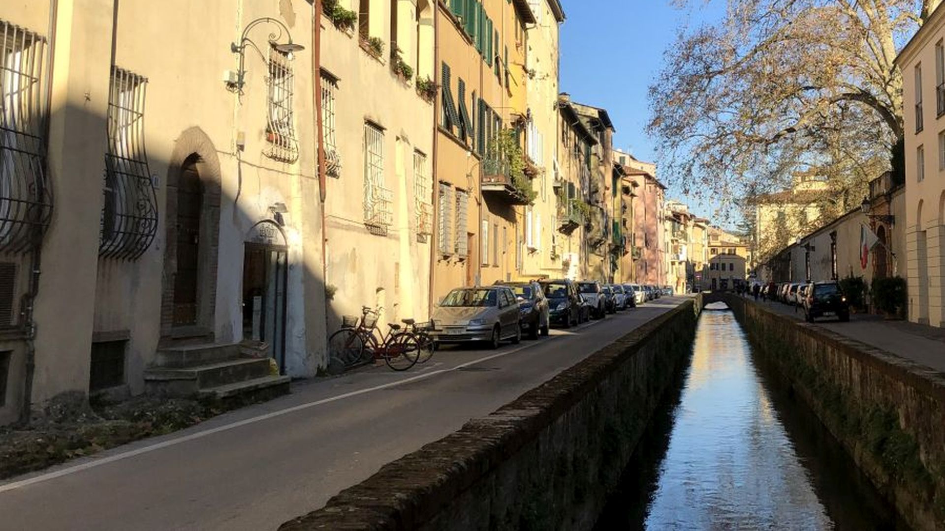 Prospettiva di via del fosso. Sulla sinistra le case con auto parcheggiate e biciclette, al centro il fosso e sulla sinistra il muro della villa Bottini da cui sporge un platano spoglio