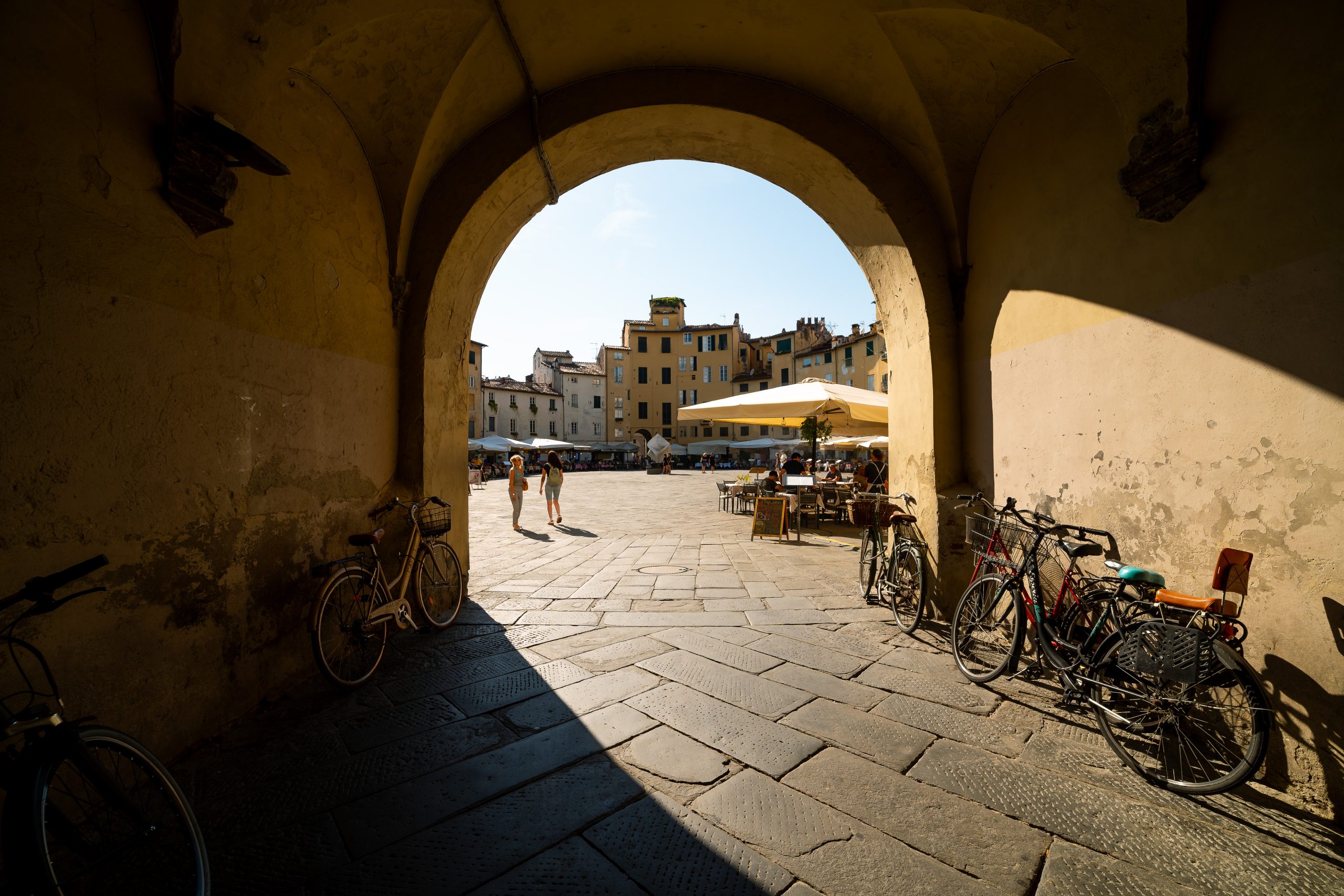 Ingresso in piazza Anfiteatro attraverso un arco. sui due lati sono appoggiate delle biciclette e nella piazza ci sono alcune persone che camminano e altre sedute ai tavolini dei bar. Sul fondo le facciate curve delle case, con tante finestre, si affacciano sulla piazza.
