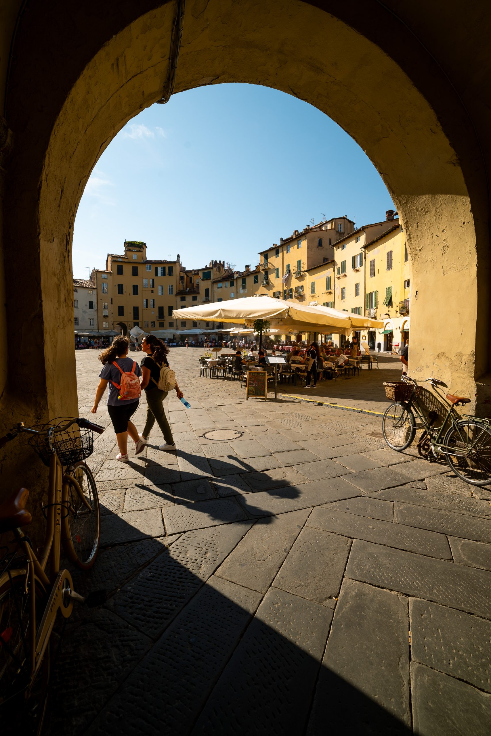 Due ragazze entrano in piazza dell'Anfiteatro di Lucca atraverso uno degli archi di ingresso. Sullo sfondo le case curve della costruzione.
