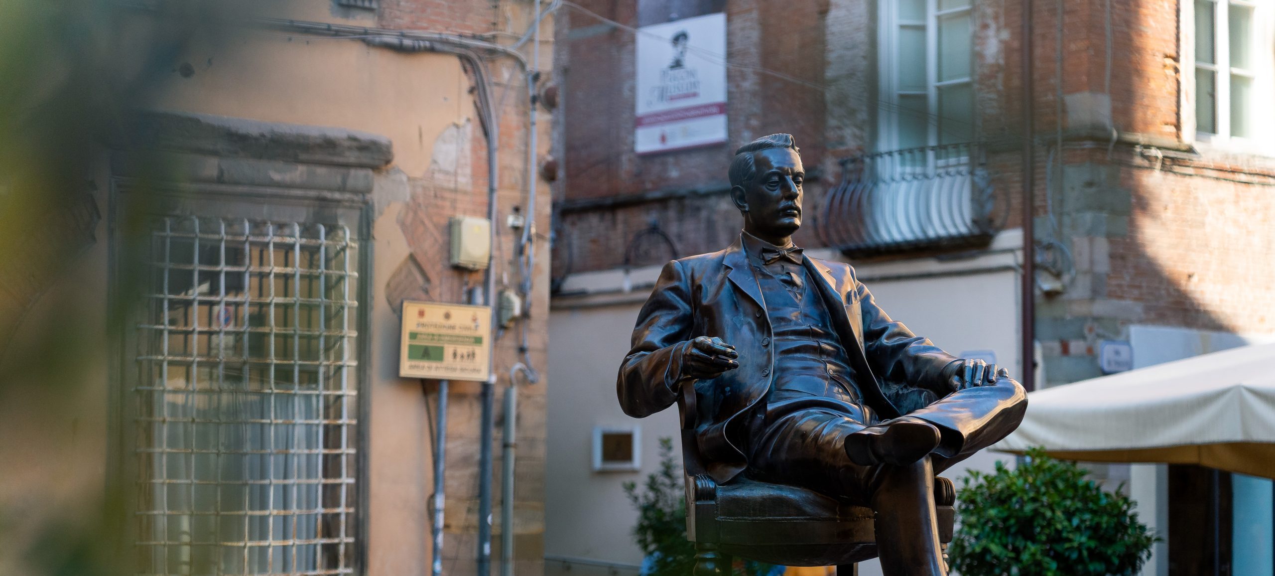 Statua di Giacomo Puccini che fuma seduto in poltrona in piazza della cittadella. Sullo sfondo la casa natale