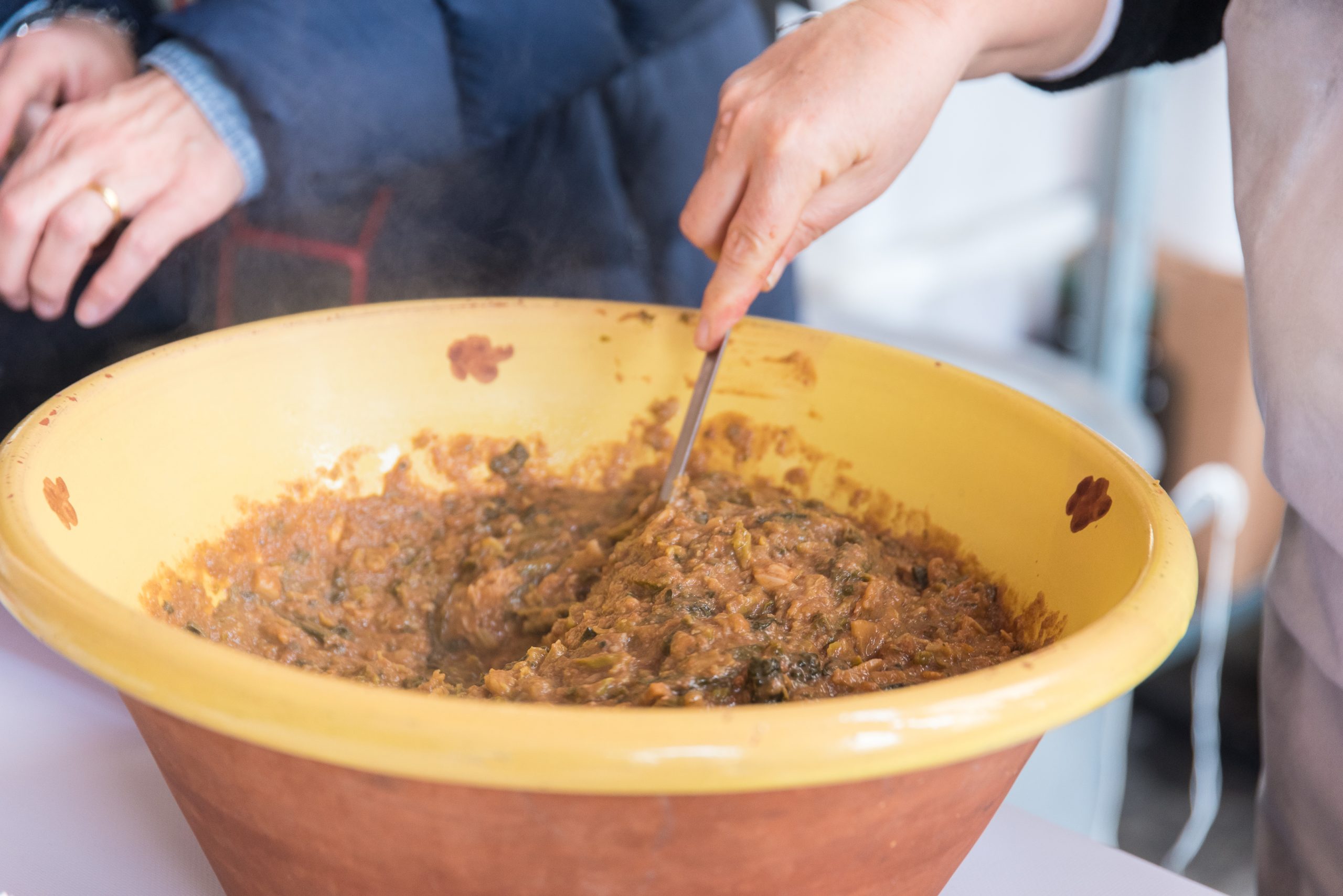 Una mano sta mescolando la zuppa alla frantoiana in una zuppiera gialla di terracotta smaltata con i motivi tipici lucchesi della