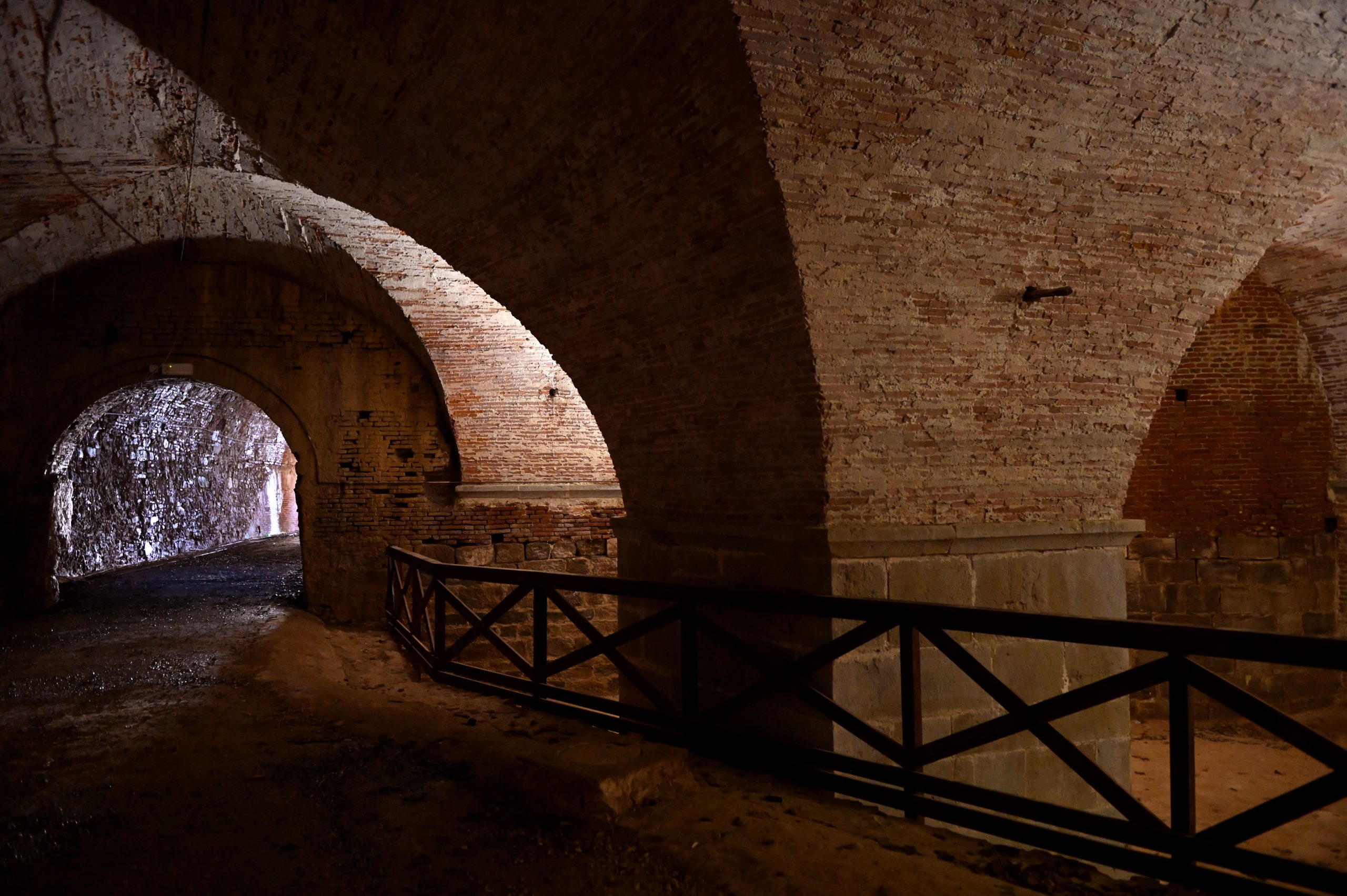 il soterraneo del baluardo San Colombano delle Mura di Lucca. Grandi pilastri rettangolari sorreggono le volte in mattoni. Dall'alto e dal fondo di una galleria che si apre sulla sinistra, filtra la luce dell'esterno,