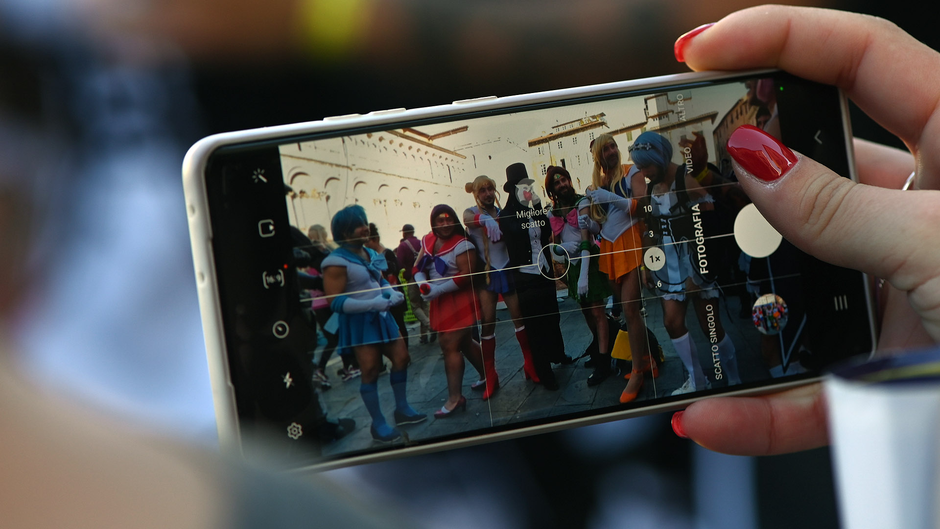 la mano di una ragazza tiene un cellulare che inquadra un gruppo di cosplayer davanti alla chiesa di san Michele a Lucca Comics & Games