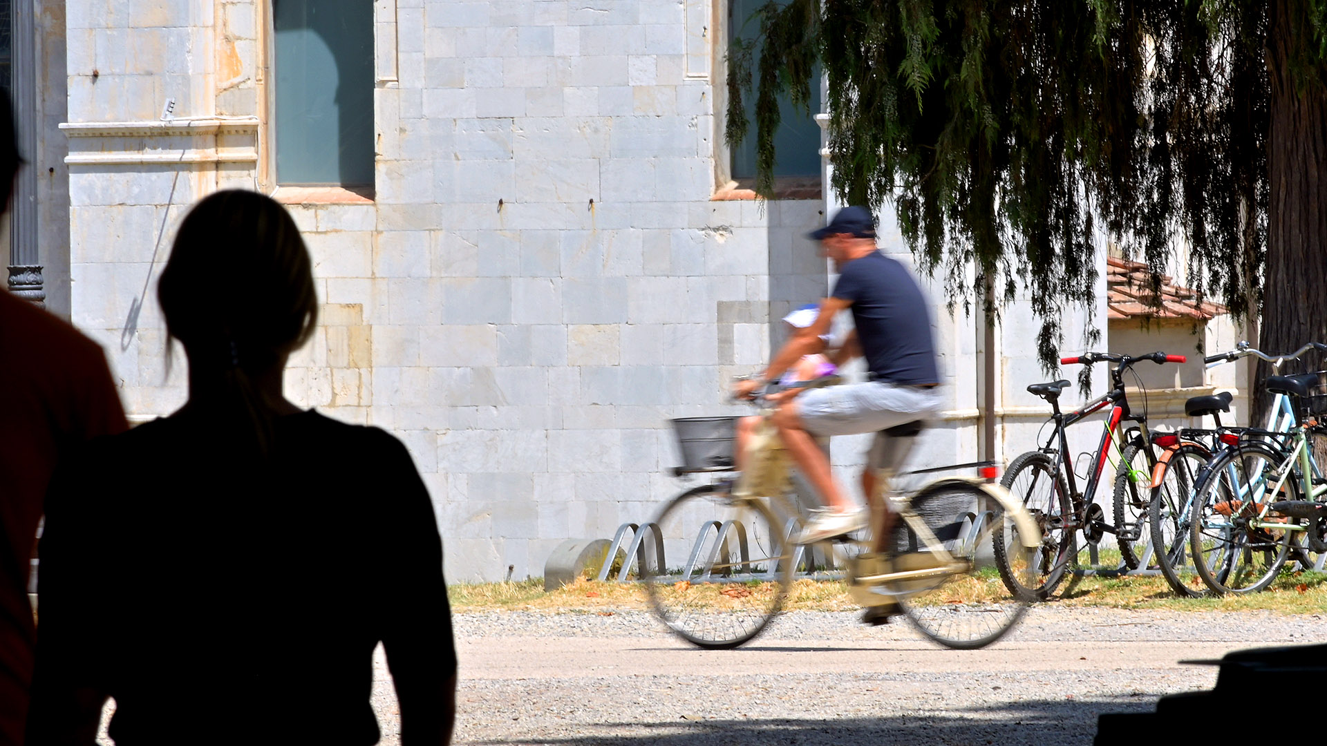 in primo piano la sagoma di una persona si staglia sul muro bianco della cattedrale. Sulla destra ci sono delle biciclette parcheggiate e una persona che attraversa la scena in bicicletta.